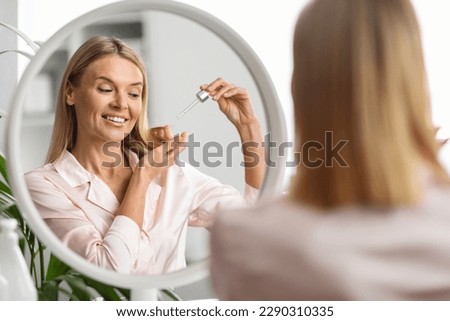 Happy Middle Aged Woman Applying Moisturising Oil On Dry Hair Ends, Smiling Mature Female Sitting Near Mirror At Dressing Table, Making Haircare Routine At Home, Selective Focus On Reflection
