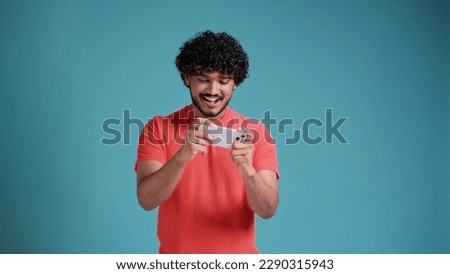 Excited happy young indian man gamer player using smartphone, playing game in mobile videogame app or watching digital video stream holding cell phone isolated on blue studio background
