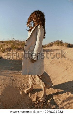 stylish woman in a light jacket posing on the sand, turning away from the camera