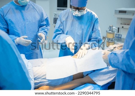Front view of a Caucasian female surgeon with team, in blue surgical uniform, face mask, and medical loupes, standing surgery on the patient's stomach through a surgical drape in the operating room. Royalty-Free Stock Photo #2290344795