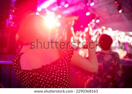 Back view of woman in polka dot dress dancing near stage during open air music festival against red glowing lamp