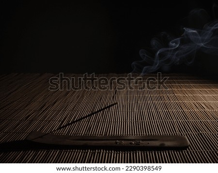 Flat wooden incense holder with an incense burning and releasing smoke on a bamboo table with a black background