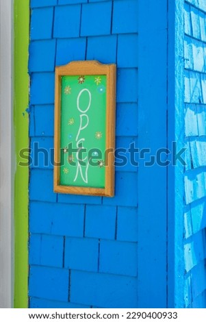 A vibrant green colored sign with small painted flowers, a wooden frame and a vibrant royal blue exterior wooden wall. The trim on the house is lime green. The boards are blue painted cedar shakes. 