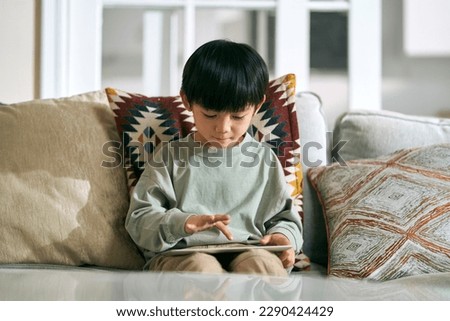 five-year-old little asian boy sitting on family couch at home using digital tablet computer Royalty-Free Stock Photo #2290424429