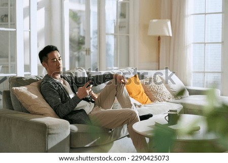 young asian adult man sitting on family couch in living room at home looking at cellphone Royalty-Free Stock Photo #2290425073