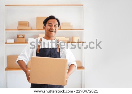 Photo of young Asian man retail seller, entrepreneur, online store drop shipping small business owner looking at camera standing in delivery shipping warehouse with parcel boxes.