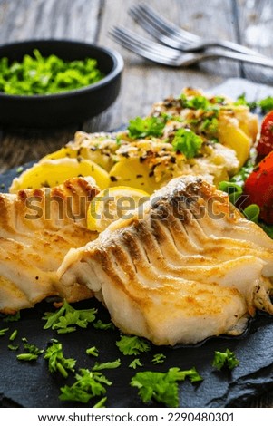 Fried cod loin with baked potatoes and fresh vegetables on black plate on wooden table 