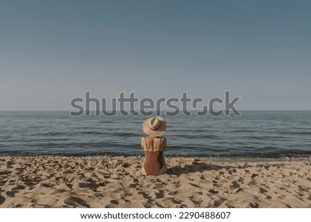 Aesthetic summer vacation fashion concept. Young tanned woman wearing a swimsuit and straw hat is sitting and relaxing on tropical beach with sand and watching at sea