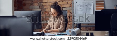 Professional female interior designer looking at blueprint while working on new design project, sitting at the table in her office
