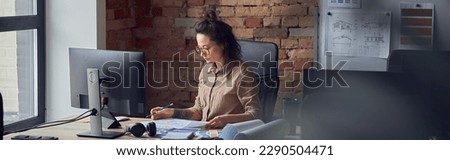 Concentrated woman, professional designer looking through papers while working on new interior design project, sitting at the table in her office