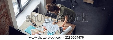 High angle view of interior designer or architect in casual wear with messy hairdo working with color samples and blueprint for new project, standing in her office