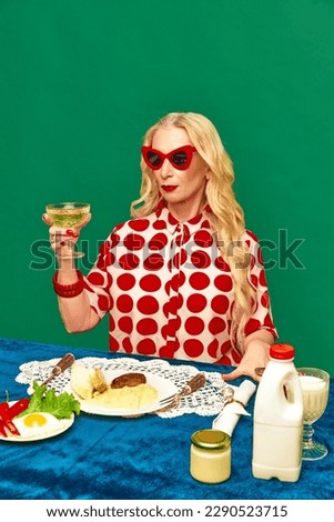 Dinner time. Attractive senior lady drinking cocktail, eating smashed potatoes and cutlet against green studio background. Food pop art photography. Complementary colors. Concept of taste, creativity