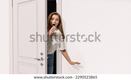 Young woman peeking out open grey door, covering mouth with hand. Looking shocked, surprised, astonished. Unexpected situation. Concept of emotions, facial expression, lifestyle, news Royalty-Free Stock Photo #2290523865