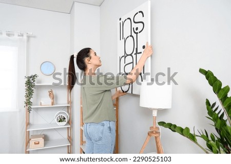 Young woman hanging picture on wall in living room Royalty-Free Stock Photo #2290525103