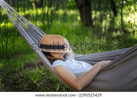Woman resting in a hammock in a summer garden covering her face with a straw hat. Summer vacation concept Royalty-Free Stock Photo #2290525429