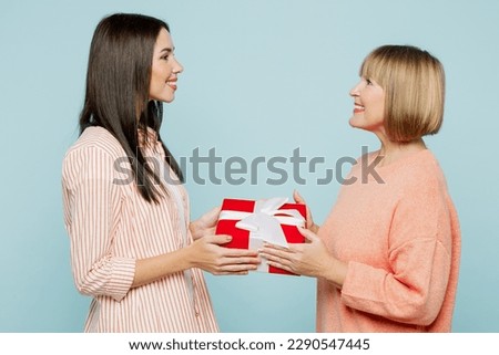 Side view fun smiling elder parent mom with young adult daughter two women together wear casual clothes hold give present box with gift ribbon bow isolated on plain blue background. Family day concept