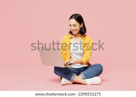 Full body young woman of Asian ethnicity wear yellow shirt white t-shirt sitting hold use work on laptop pc computer isolated on plain pastel light pink background studio portrait. Lifestyle concept