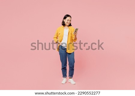 Full body young woman of Asian ethnicity wear yellow shirt white t-shirt stand hold in hand use mobile cell phone chatting browsing internet isolated on plain pastel pink background studio portrait Royalty-Free Stock Photo #2290552277