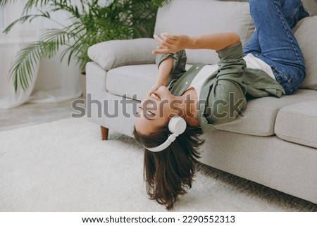 Young woman of Asian ethnicity wearing casual clothes headphones listening to music close eyes sing lay down on grey sofa couch stay at home hotel flat rest relax spend free spare time in living room