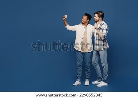 Full body young two friends happy men 20s wear white casual shirts together doing selfie shot on mobile cell phone show v-sign isolated plain dark royal navy blue background. People lifestyle concept