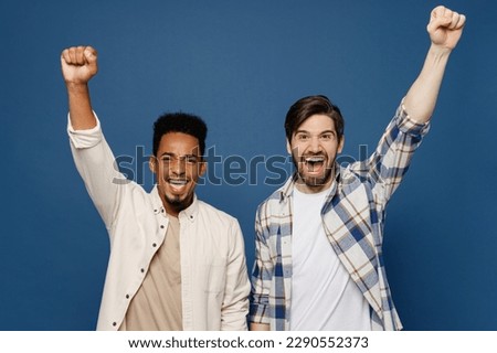 Young two friends overjoyed fun men wear white casual shirts together do winner gesture celebrate clenching fists say yes shout isolated plain dark royal navy blue background. People lifestyle concept