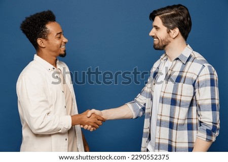 Side view young two friends cheerful men 20s wear white casual shirts looking camera together shaking hands meeting each other isolated plain dark royal navy blue background. People lifestyle concept