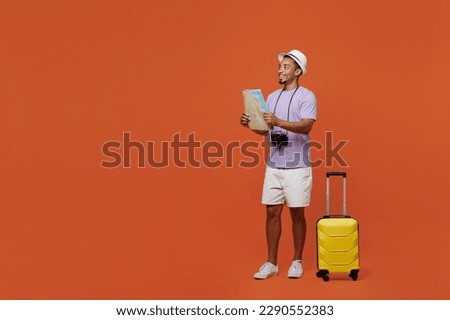 Full body traveler happy black man wear purple t-shirt hat hold suitcase read map isolated on plain orange background Tourist travel abroad in spare time rest getaway Air flight trip journey concept