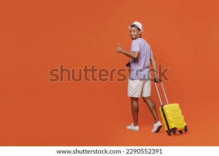 Full body traveler black man wear purple t-shirt hat go with suitcase show thumb up isolated on plain orange background Tourist travel abroad in spare time rest getaway Air flight trip journey concept Royalty-Free Stock Photo #2290552391