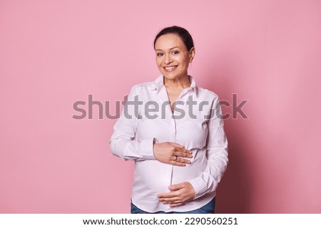 Middle aged multi ethnic woman 35-39 years , beautiful pregnant brunette holding hands on her belly, expecting baby, posing with a happy and cool smile on face, pink background. Pregnancy Maternity
