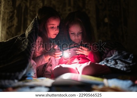 Two sisters watching movie or cartoon in smartphone together with blanket cover in bed at night home.