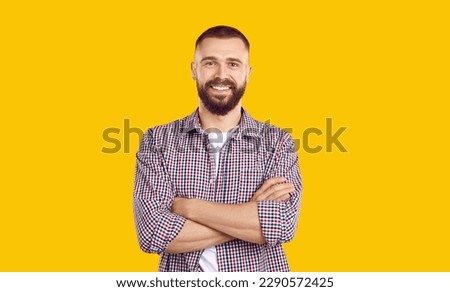 Studio portrait of a young man with arms folded. Happy handsome bearded young guy wearing a casual shirt and Tshirt standing isolated on a yellow color background, looking at the camera and smiling