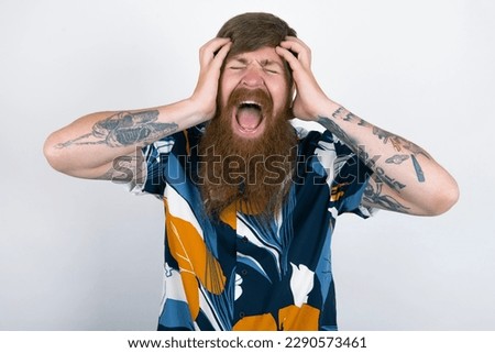 Shocked panic red haired man wearing printed shirt over white studio background holding hands on head and screaming in despair and frustration.