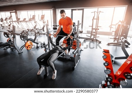 Young sporty girl works with barbell performing bench press. Personal trainer helps to perform the exercise. Wide angle shooting. Royalty-Free Stock Photo #2290580377