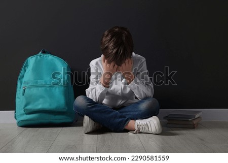 Upset boy with backpack sitting on floor near black wall. School bullying Royalty-Free Stock Photo #2290581559