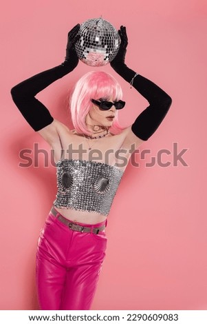 Well dressed drag queen in gloves and sunglasses holding disco ball on pink background