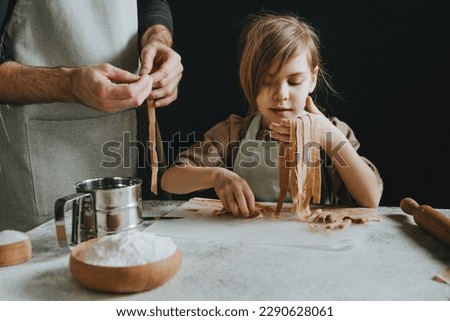 Unrecognizable father and daughter in aprons holding dough cut into strips on a white table against a dark wall. Parent with child preparing homemade pastries or noodles. Selective focus.