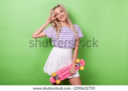 Photo of cheerful girl curly hairdo wear striped t-shirt holding skate board showing v-sign on eye isolated on green color background
