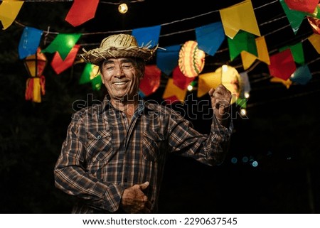 Senior man celebrating the Brazilian Festa Junina. Portrait of a man wearing typical clothes and a straw hat under the decoration of pennants and lights from the traditional June festival in Brazil. Royalty-Free Stock Photo #2290637545