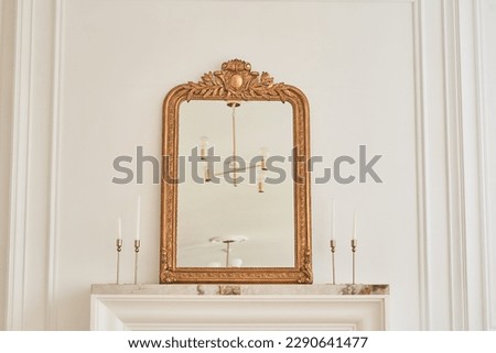 A mirror in a classic frame on the mantelpiece next to candlesticks with candles. Part of the interior with a reflection of the chandelier on the ceiling. Classic interior with copy space. 