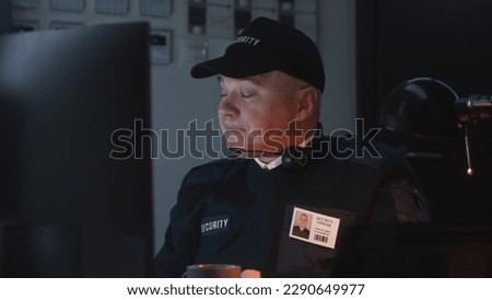 Tired security officer sits on workplace and looks view from security cameras displayed on monitor. Man drinks cup of coffee or tea. Monitoring system and CCTV. Late night working. Portrait view.