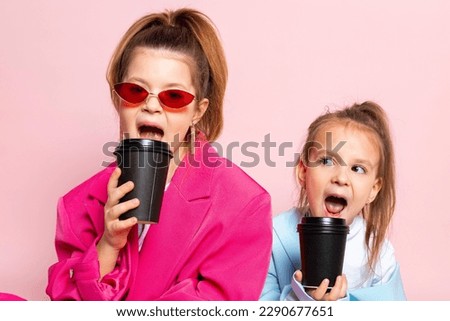 Two little girls dressed like adults. Children wearing oversized clothes. fashionable kids are holding paper cups. Isolated on pink background. Royalty-Free Stock Photo #2290677651