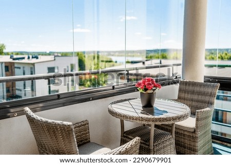 Balcony in summer. Home patio with a view. Condo apartment in city. Scandinavian decor and furniture design. Glass windows, glazing. Flower or plant on table and chairs. Sunny terrace in modern flat. Royalty-Free Stock Photo #2290679901