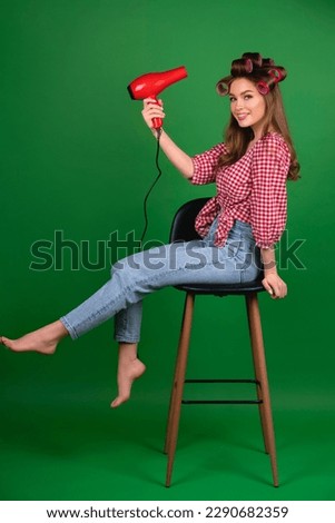 Pretty young smiling girl in big red curlers drying hair with red dryer. Studio work with attractive model in checkered red shirt and jeans sitting on chair, posing  on green background isolated.