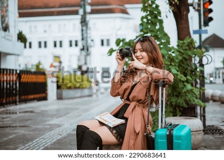 beautiful traveller in brown trench coat taking picture with her camera while sitting on the stone seat at the sidewalk