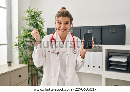 Young woman wearing doctor uniform holding smartphone screaming proud, celebrating victory and success very excited with raised arm  Royalty-Free Stock Photo #2290686017