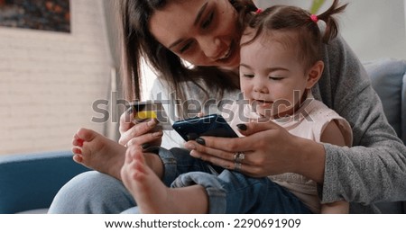 Relaxed caucasian mom is making an online purchase with credit card using smartphone while holding her cute little baby girl on knees - happy family, technology, finance 