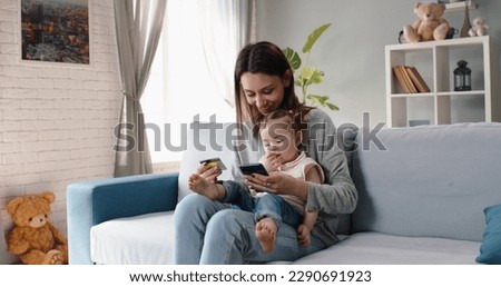Relaxed caucasian mom is making an online purchase with credit card using smartphone while holding her cute little baby girl on knees - happy family, technology, finance 