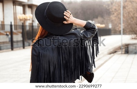 street style photo, a girl in a black outfit, in a hat and a fringed leather jacket, view from the back Royalty-Free Stock Photo #2290699277