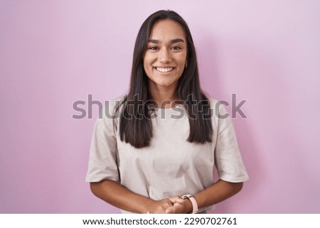 Young hispanic woman standing over pink background with hands together and crossed fingers smiling relaxed and cheerful. success and optimistic 