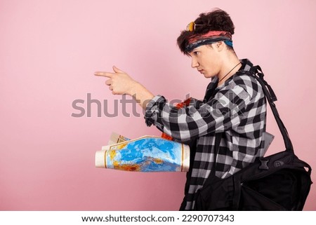A young handsome traveller or tourist carrying the travelling equipment and looking at the map. Isolated on pink background.